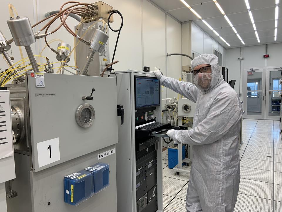 A person in a white coverall and safety glasses poses standing next to a rack of computers and other devices in a lab. 