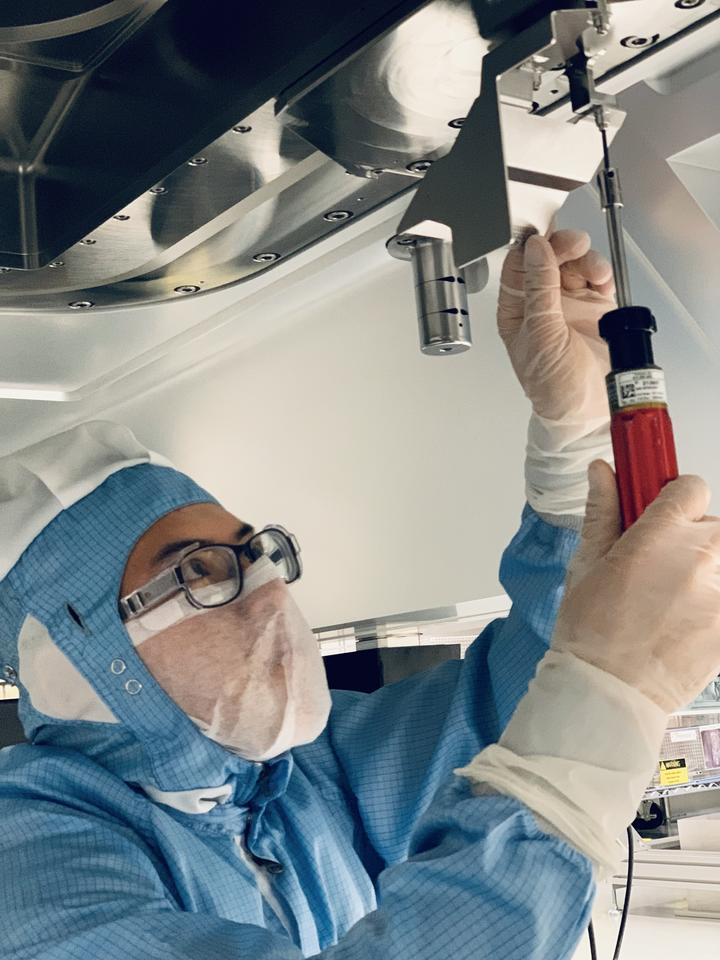 A person in a blue coverall and safety glasses reaches up to work on the underside of a large scientific device. 