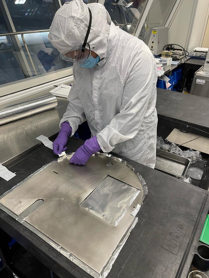 A person in a white coverall and safety glasses bends over a table where she is working on a flat piece of metal. 