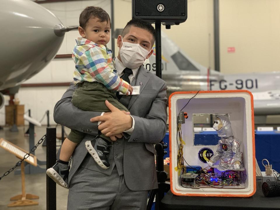 A man in a suit holds a toddler and poses next to a box containing wiring and a toy monkey in a space suit, in a large museum space with aircraft in the background. 