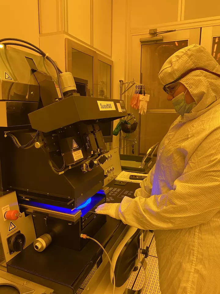 A person in white coveralls and safety glasses looks down at a machine she is using in a lab with orange lighting. 