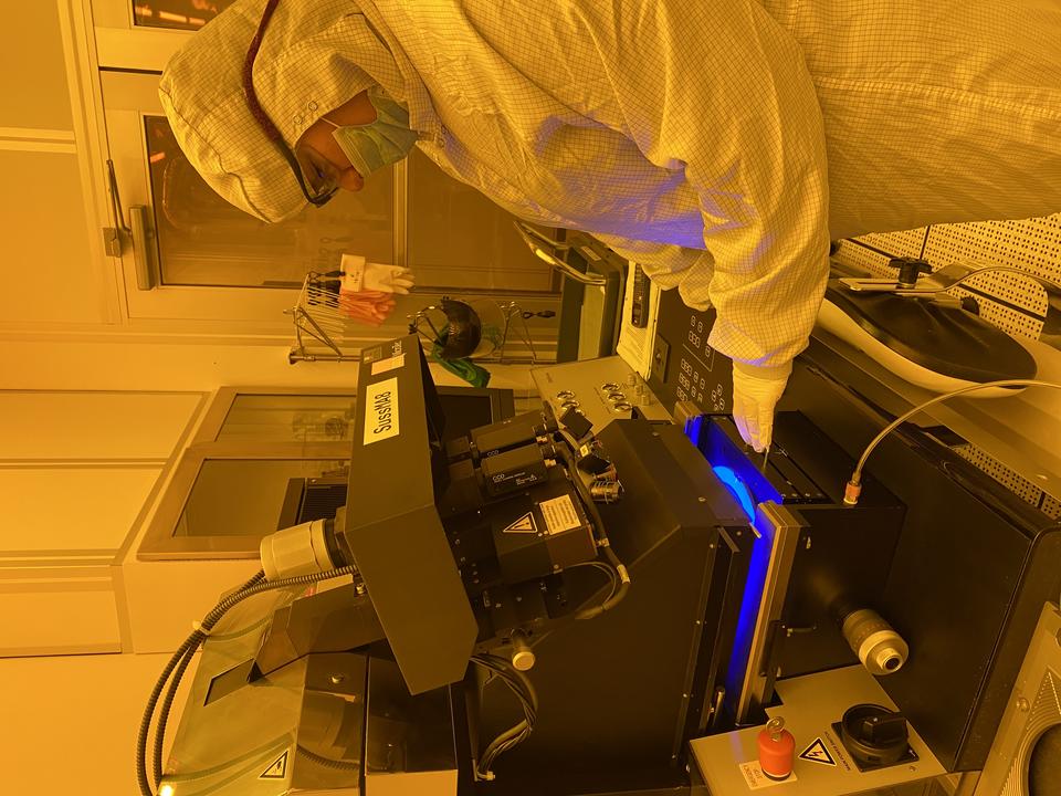 A person in white coveralls and safety glasses looks down at a machine she is using in a lab with orange lighting. 