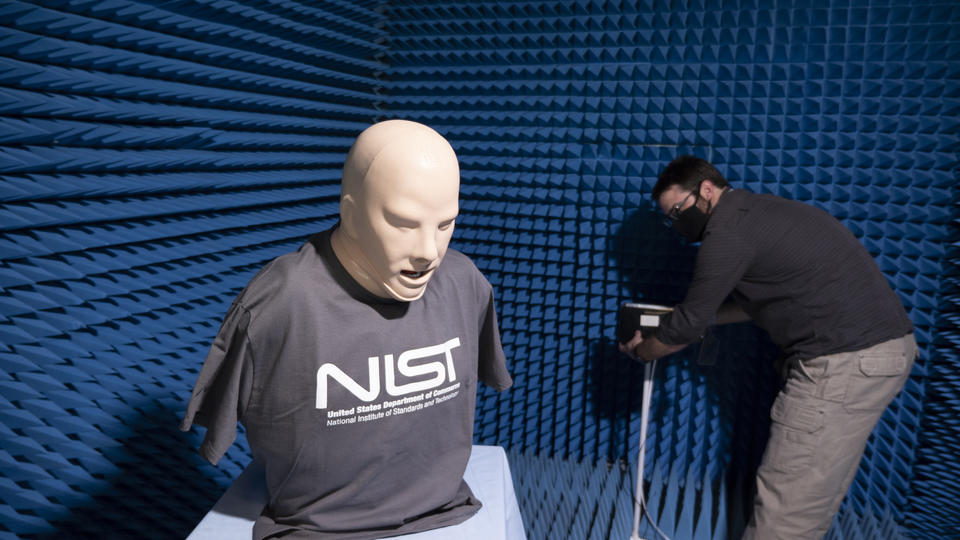 A model head and torso wearing a NIST T-shirt stands on a table in a small room with blue foam on the walls, while a researcher sets up equipment in the background.