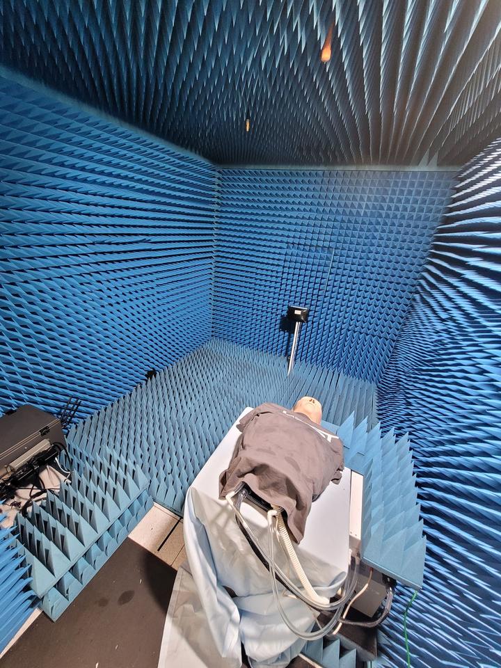 A model head and torso wearing a gray T-shirt lies on a table in a small room with blue foam on the walls. Tubes run from the torso to equipment beneath.