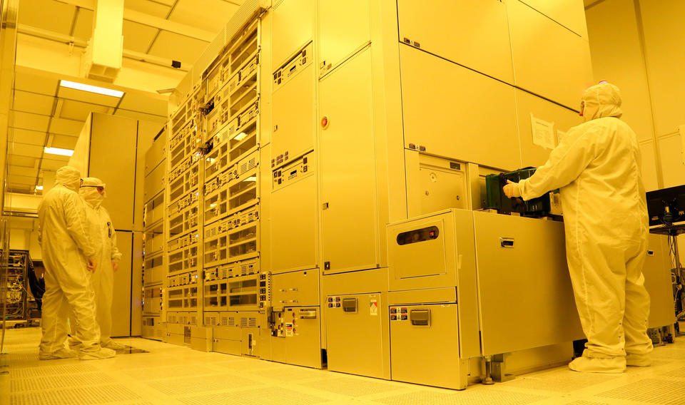 Three individuals in white suits that cover their full bodies from head to toe stand at a large, floor-to-ceiling machine with panels and glass windows, in a room with yellow lighting.