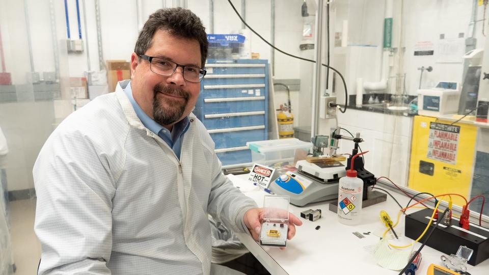 John Kitching poses at a lab table, displaying a chip-scale device in a clear plastic box. 