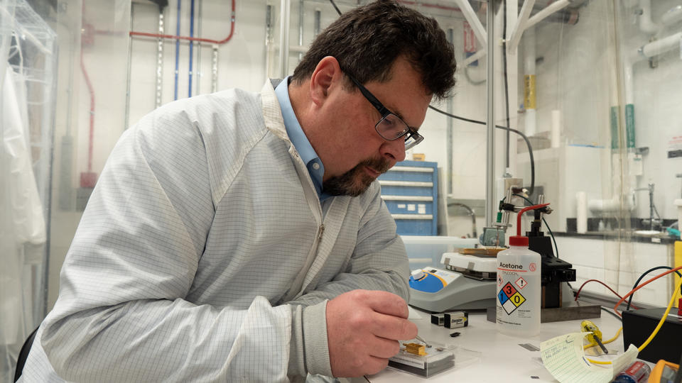 John Kitching leans over a table in the lab, using tweezers to point to part of a chip-scale device in a clear plastic box. 