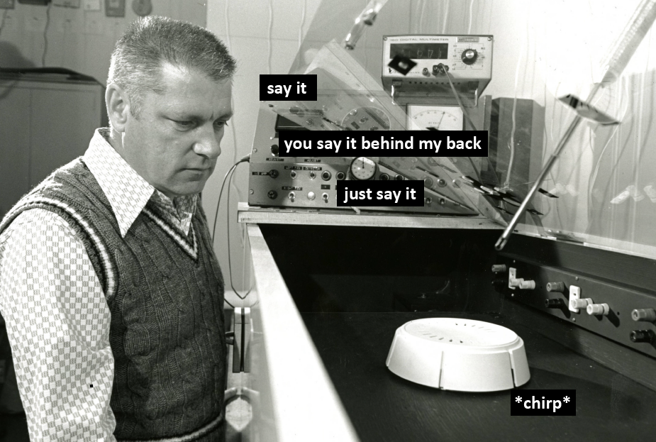 A man looks down at a device on a table. Text reads: "Say it. You say it behind my back. Just say it." Text alongside the smoke detector device reads: "Chirp."