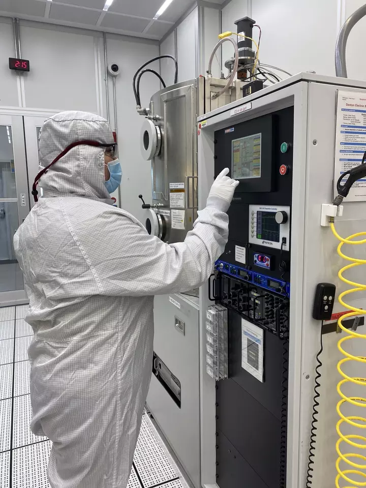 A person in a white coverall and mask stands at a screen on a large piece of equipment in a lab. 