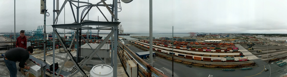 Wide-angle photo shows two people on the left installing equipment on the roof of a building, with a view of a port and rows of shipping containers to the right. 