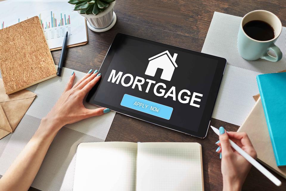 View looking down at a desk shows coffee, books and hands holding a tablet with the screen saying "Mortgage: Apply Now."