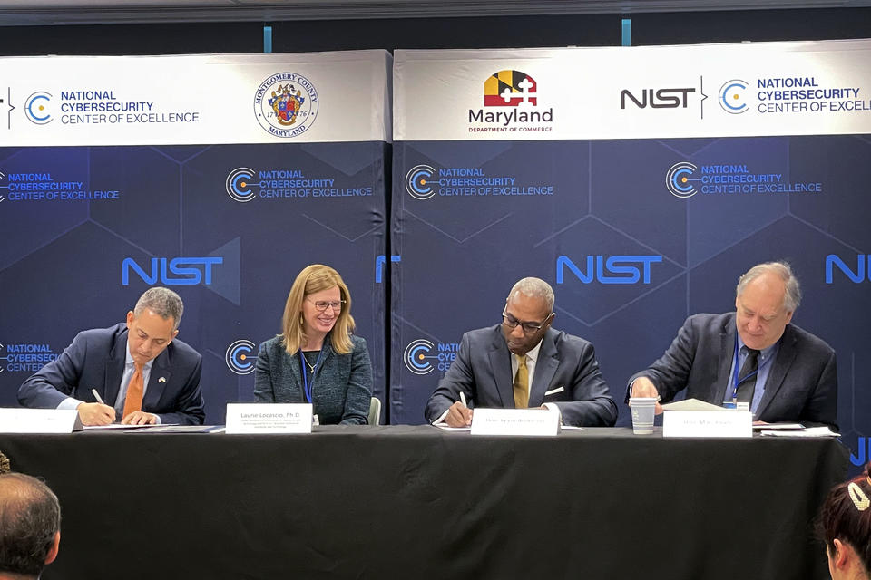 Four officials sit at a table signing documents in front of banners for the NIST National Cybersecurity Center of Excellence.