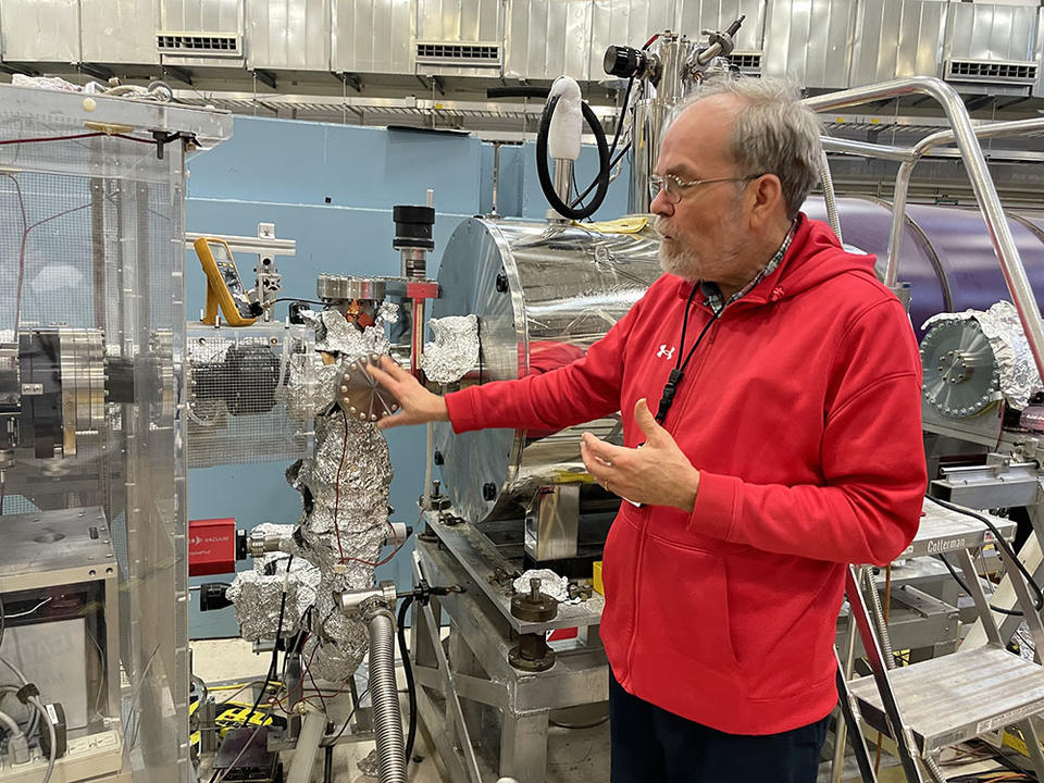 Scott Dewey stands and gestures toward the end of a long metal tube where many connectors and devices are wrapped in aluminum foil. 