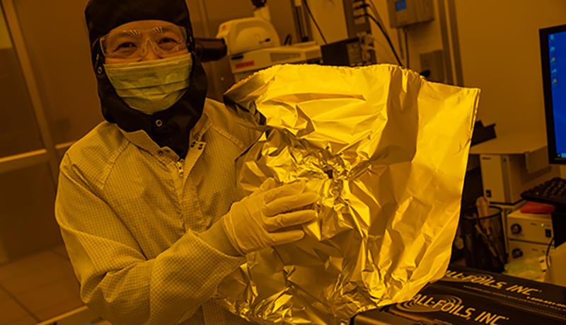 NIST researcher Jessie Zhang, wearing a protective suit, mask, and safety glasses holds up a piece of foil in the lab. 