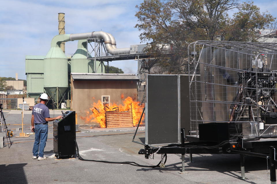 A photo shows an outdoor experimental setup with a huge fan blowing fire toward a shed. 