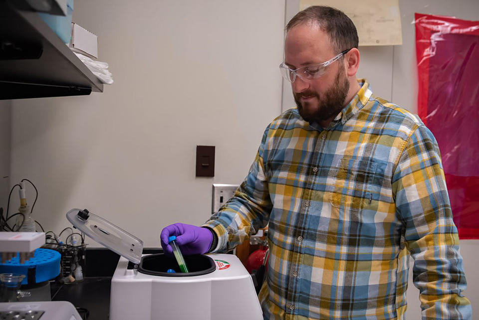 Ben Place, wearning safety glasses and gloves, places a vial into a circular opening on a tabletop centrifuge machine. 