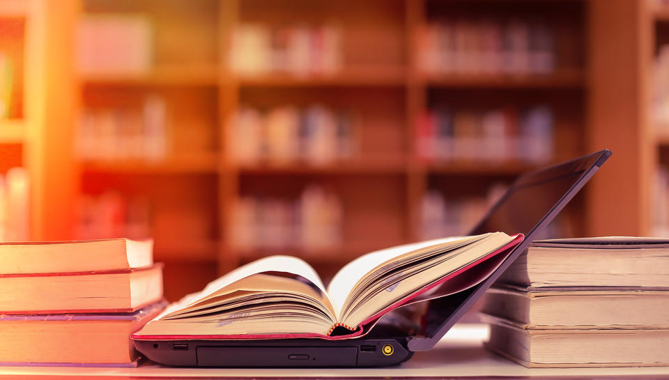 An open book sitting on top of an open laptop surrounded by stacks of books and bookshelves