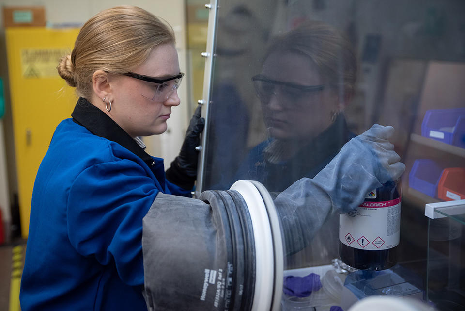 Christine McGinn wears safety glasses as she opens a container inside a clear plastic box in the lab. 