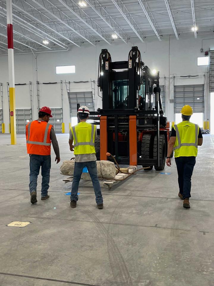 Three people in hard hats and other safety gear stand in front of a forklift carrying a piece of concrete inside a warehouse.