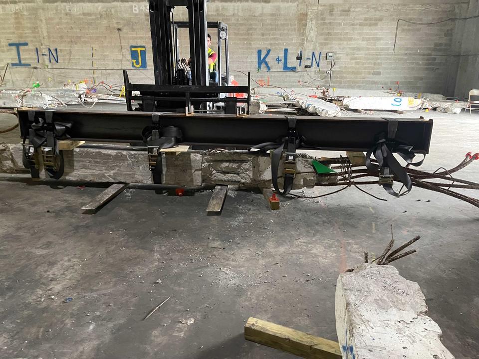 Inside a warehouse, a steel beam is attached to a concrete column being lifted by a forklift.