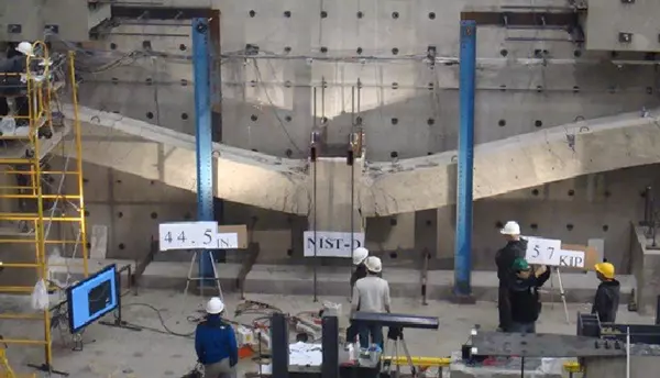 Researchers in hard hats stand inside a large indoor space studying a horizontal concrete test structure on a wall.