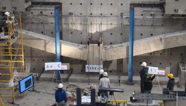 Researchers in hard hats stand inside a large indoor space studying a horizontal concrete test structure on a wall.