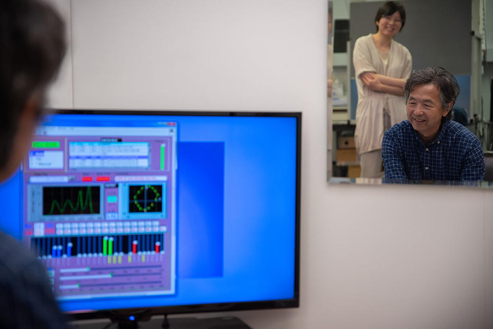 A mirror reflects Yoshi Ohno, seated, and Jane Li, standing, as they view a computer screen showing lighting data. 