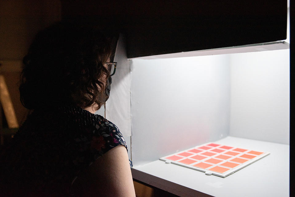 Megan King looks into a lighting test booth at a grid of orange-hued color samples. 