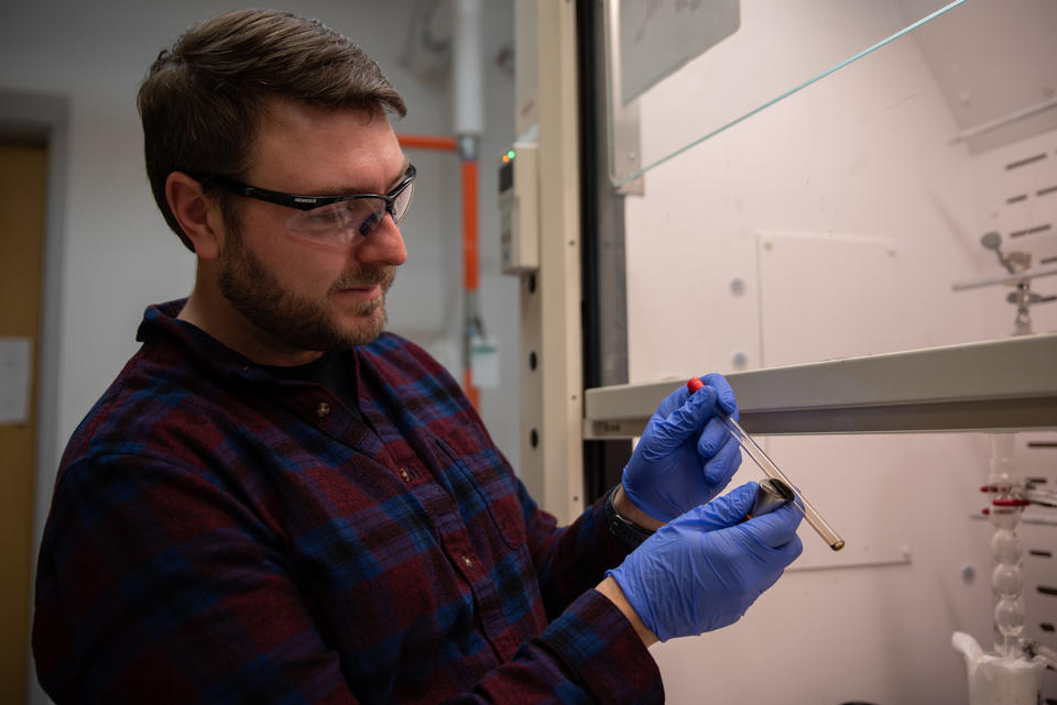 A man wearing safety goggles and rubber gloves inspects a test tube he is holding alongside a magnet. 