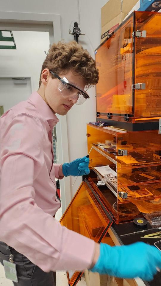 Win Lawson wears safety glasses as he stands at a lab table with an open cabinet made of translucent orange-tinted plastic.