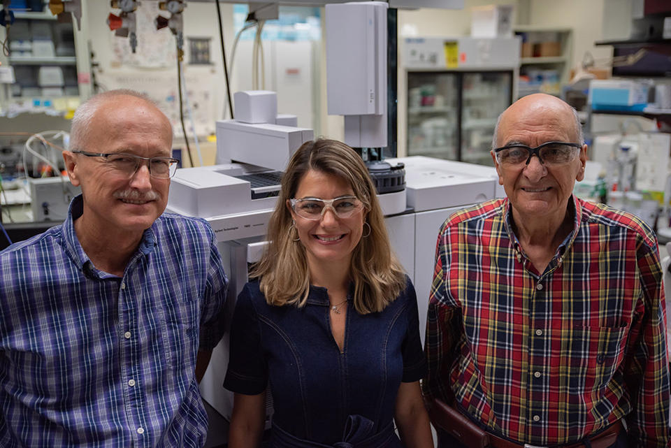 Three researchers pose smiling in the lab, standing in front of a large piece of equipment.  
