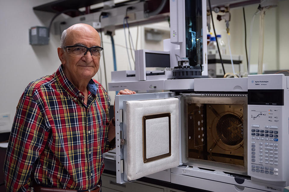 Miral Dizdaroglu wears safety glasses as he poses in the lab next to an open spectrometer, which looks a little like a microwave with extra buttons. 