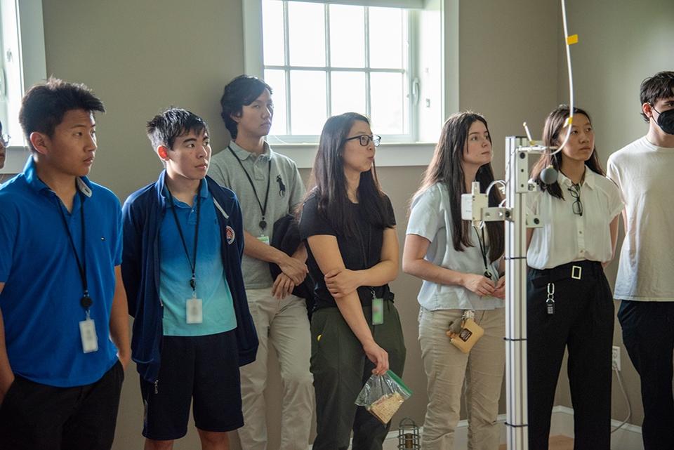 Half a dozen high school students stand listening to a presentation in a room with scientific equipment.