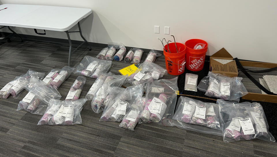 Packets of cylindrical objects wrapped in clear plastic bags with white labels are lined up on the floor of a conference room.