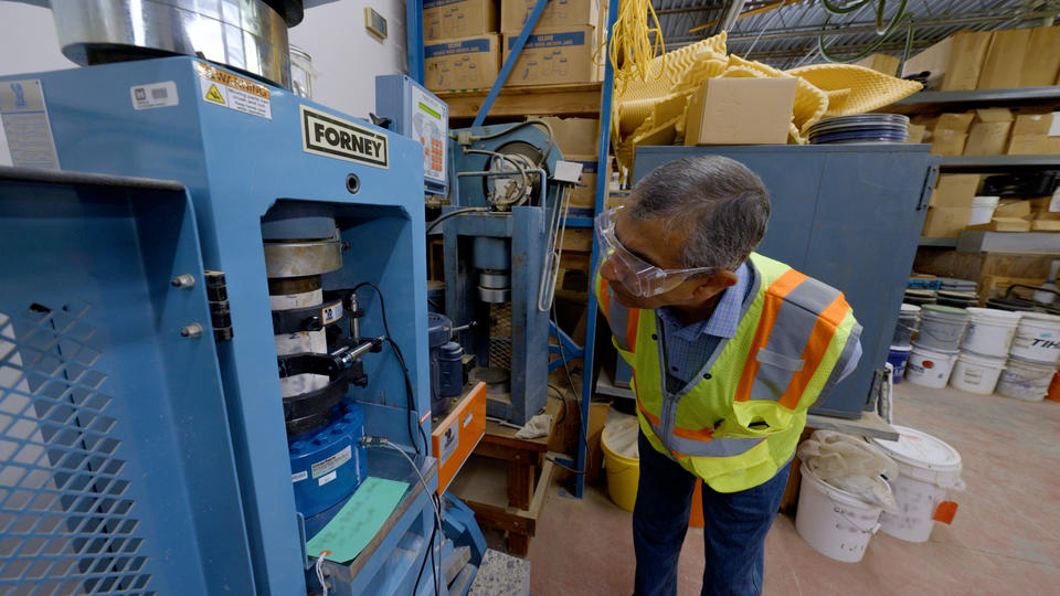 A researcher in safety gear leans over to examine a concrete core sample placed inside a blue metal testing machine.