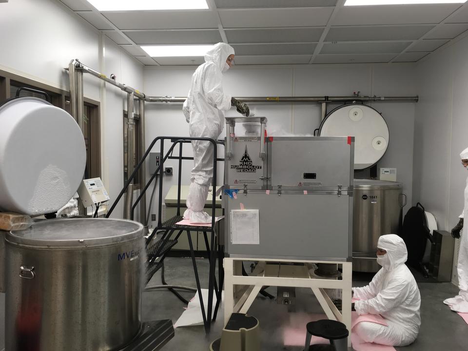 A researcher in a white coverall stands on a stool feeding material into a large square machine putting off cold vapor, while another researcher sits at the bottom to collect material coming out. 