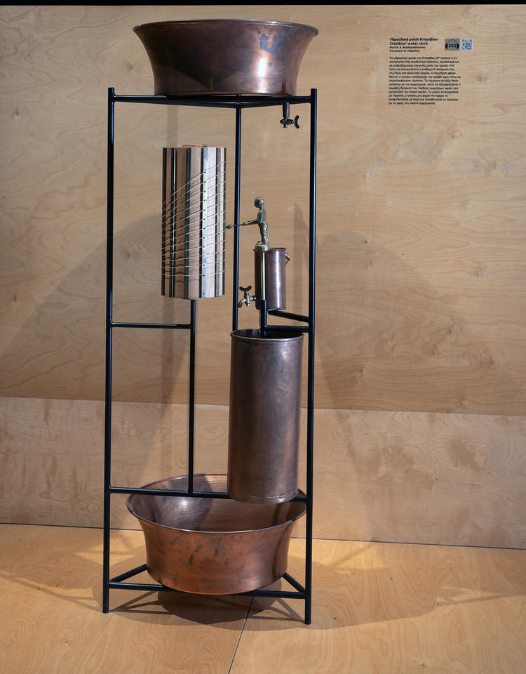 Ancient water clock is a vertical device with a copper-colored pot on top, valves and pipes leading down, and another pot at the bottom. 