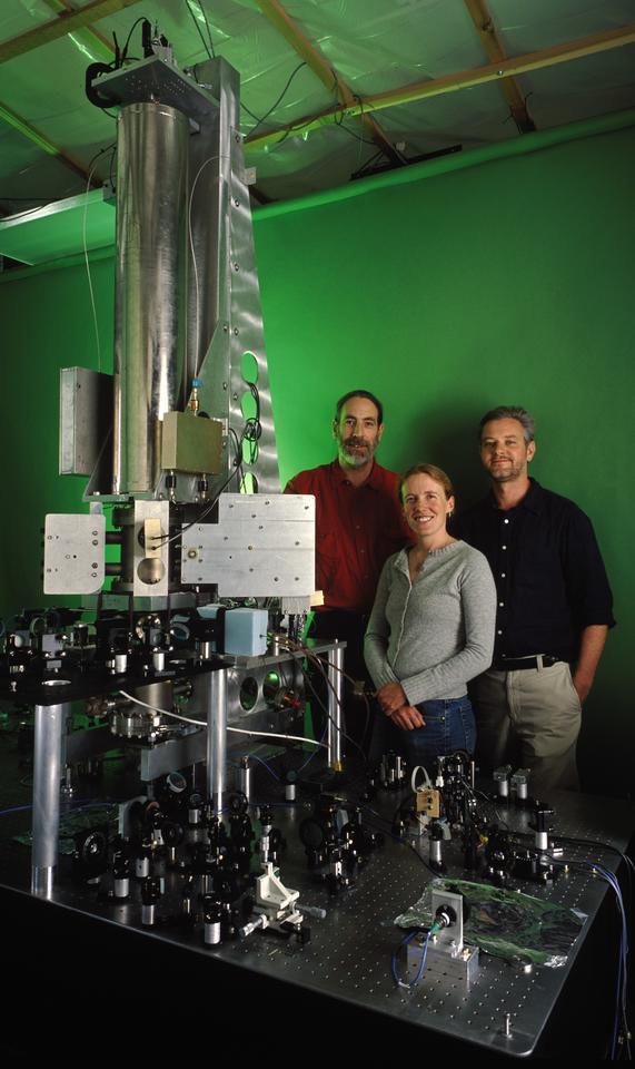 Two men and a woman pose standing behind a large scientific device with a metal cylinder reaching to the ceiling and green light shining out. 