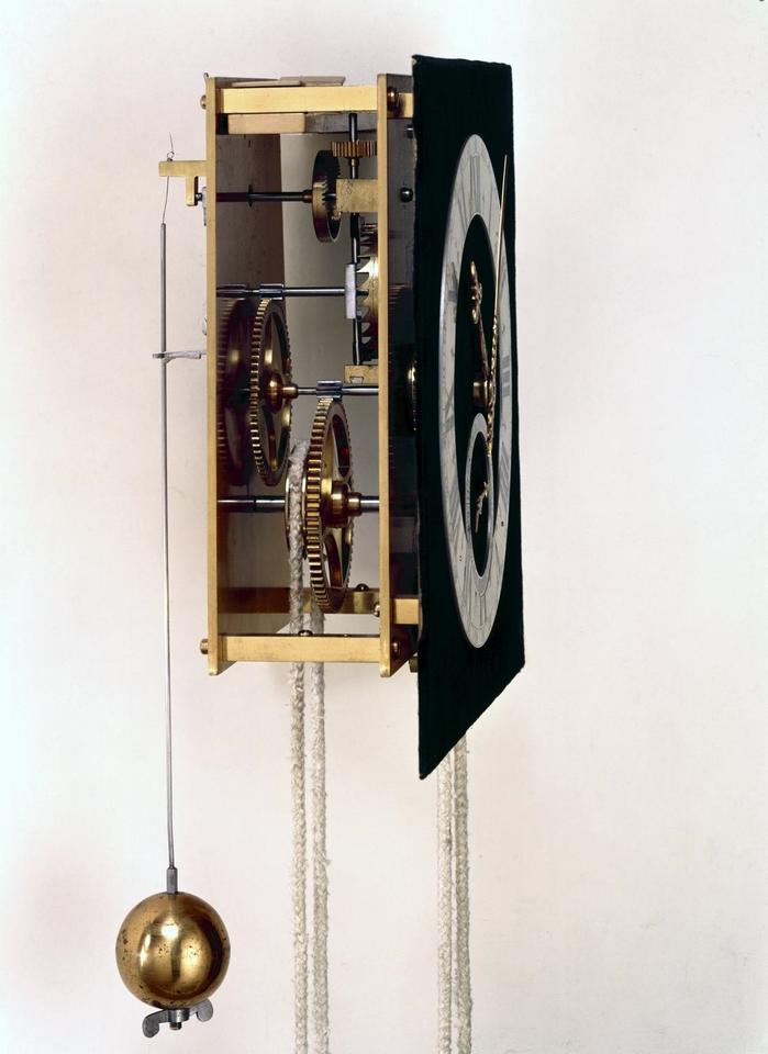 Side view of antique clock shows the gears behind the clock face and a spherical pendulum hanging from a wire. 