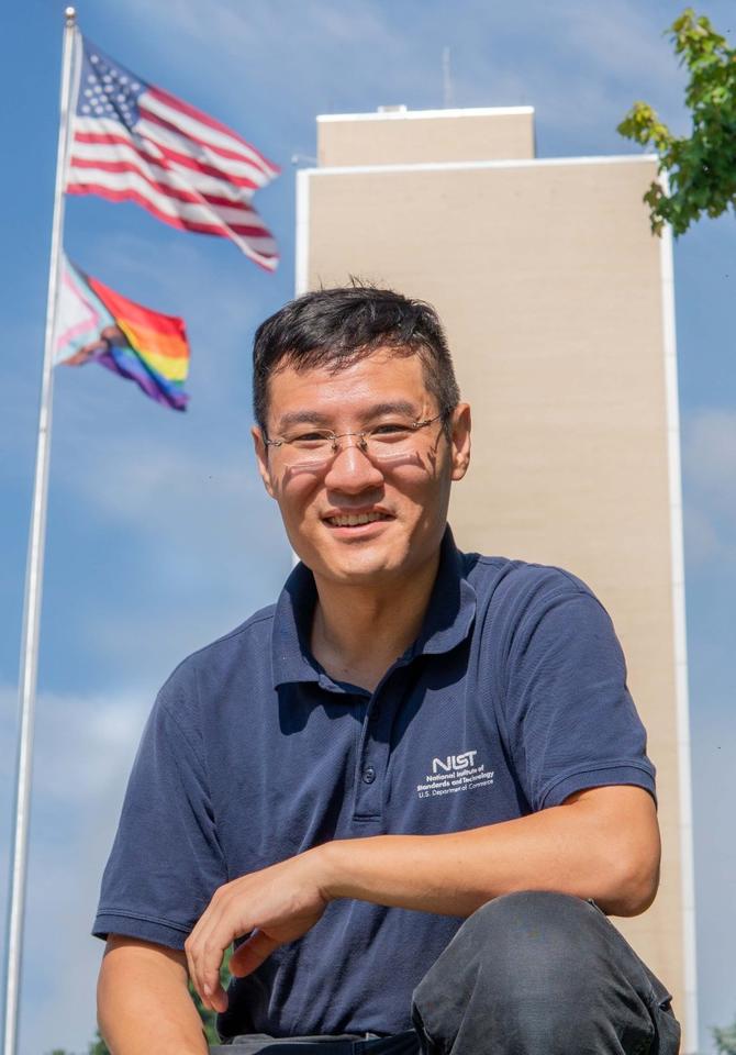 Dr Raymond Sheh in front of Building 101 and main flagpole, flying the US and Progressive Pride flags, at the main NIST campus in Gaithersburg. 