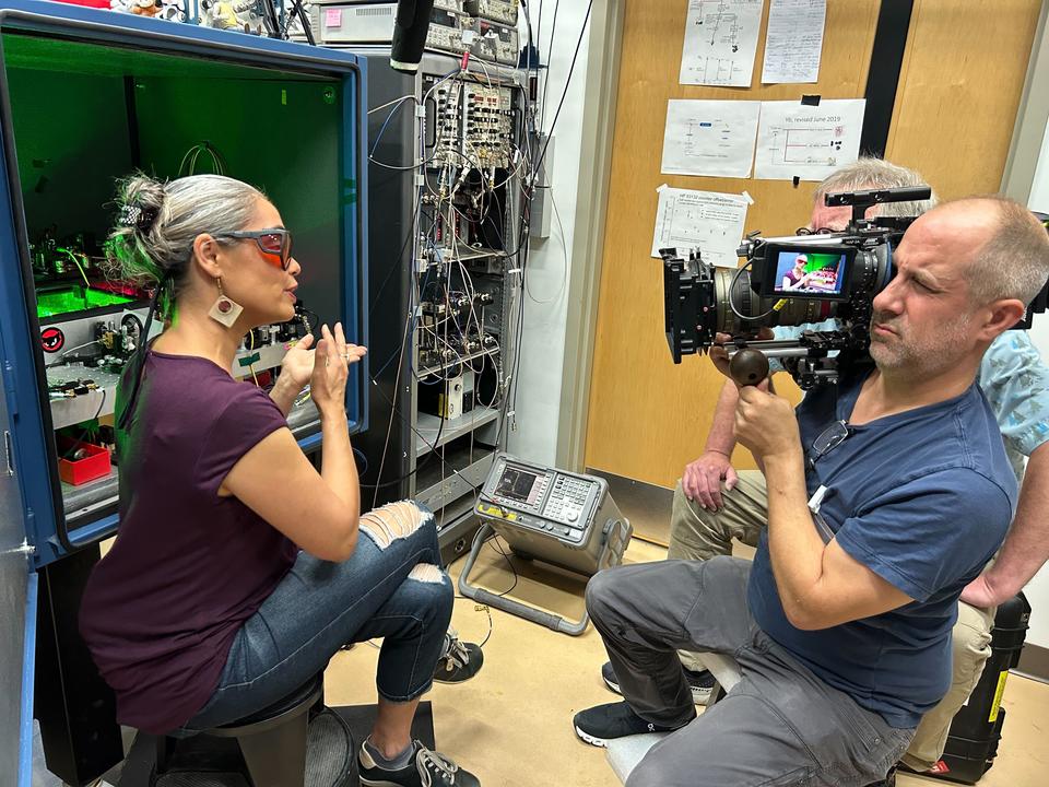 A woman in safety glasses sits on a stool in the lab, speaking and gesturing while being filmed by a man holding a video camera. 