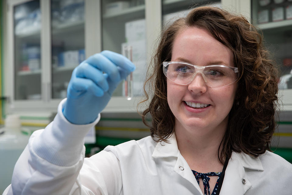 Jerilyn Izac, wearing safety glasses and a lab coat, holds up test tube containing clear liquid.