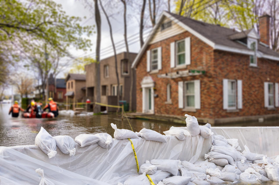 Flood protection sandbags buffer waters around flooded homes with rescue team pushing boat through flooded streets.