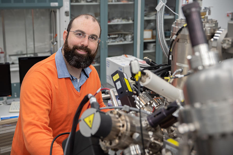 Trey Diulus poses sitting in the lab with a large, complex scientific instrument in the foreground.