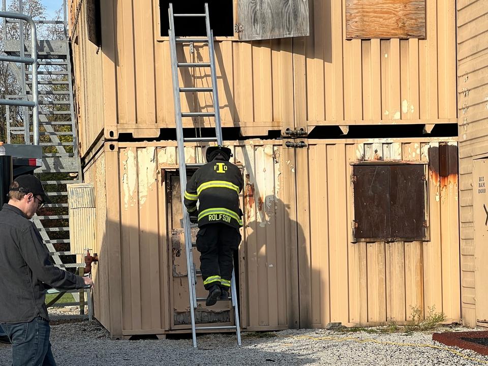 A firefighter climbs a ladder into a shipping container, while a person to their left tracks their location.