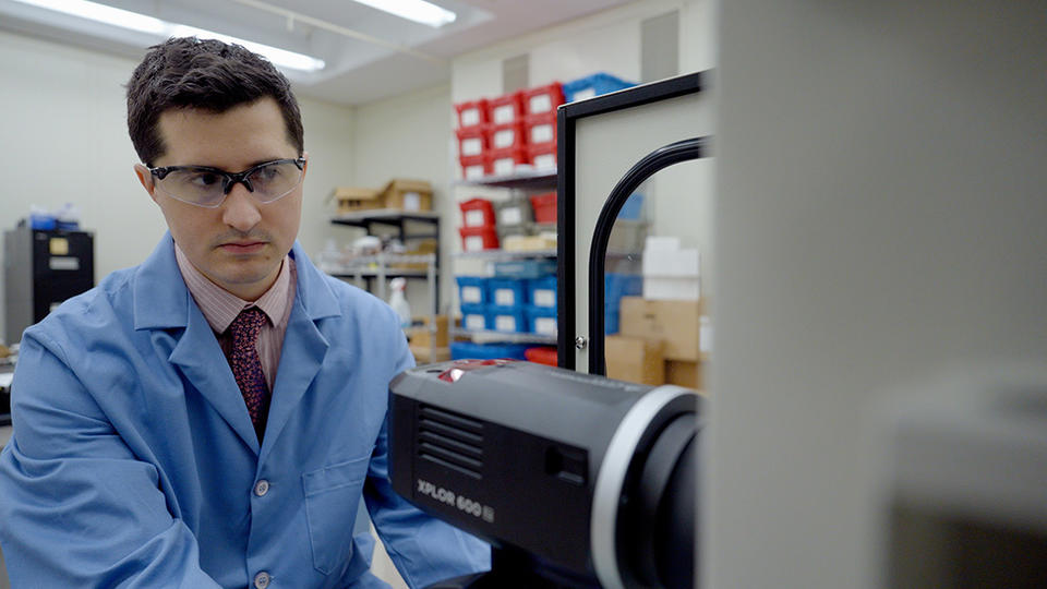 Ryan Falkenstein-Smith, wearing a lab coat and safety glasses, adjusts a black camera-like device in the lab. 