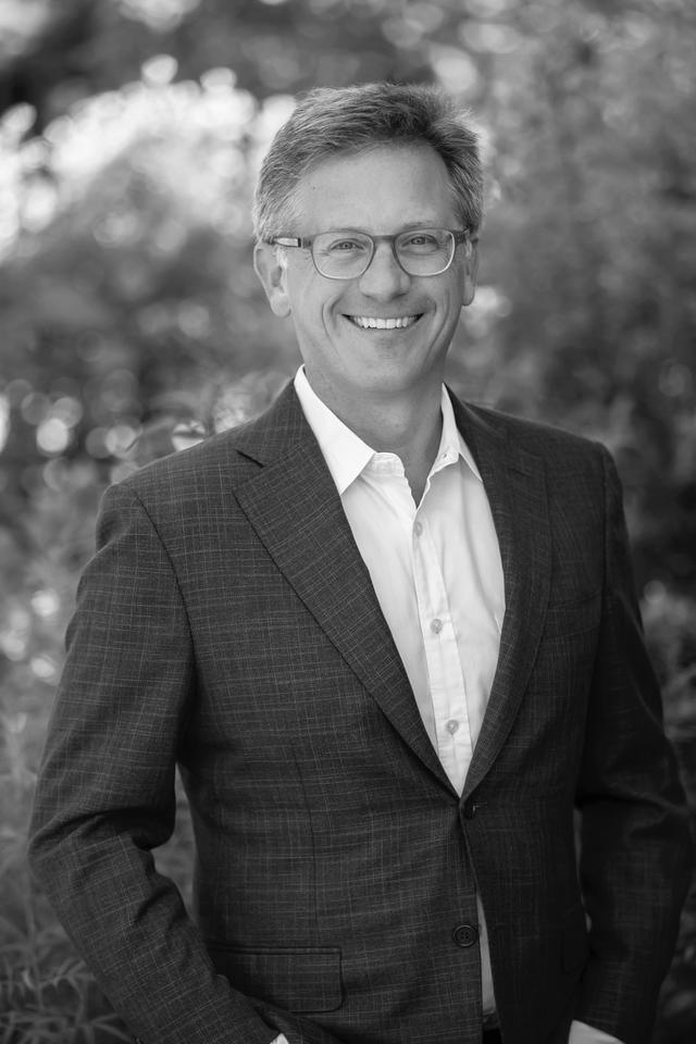 black and white picture of a man with glasses, dark suit coat and white collared shirt. Natural background