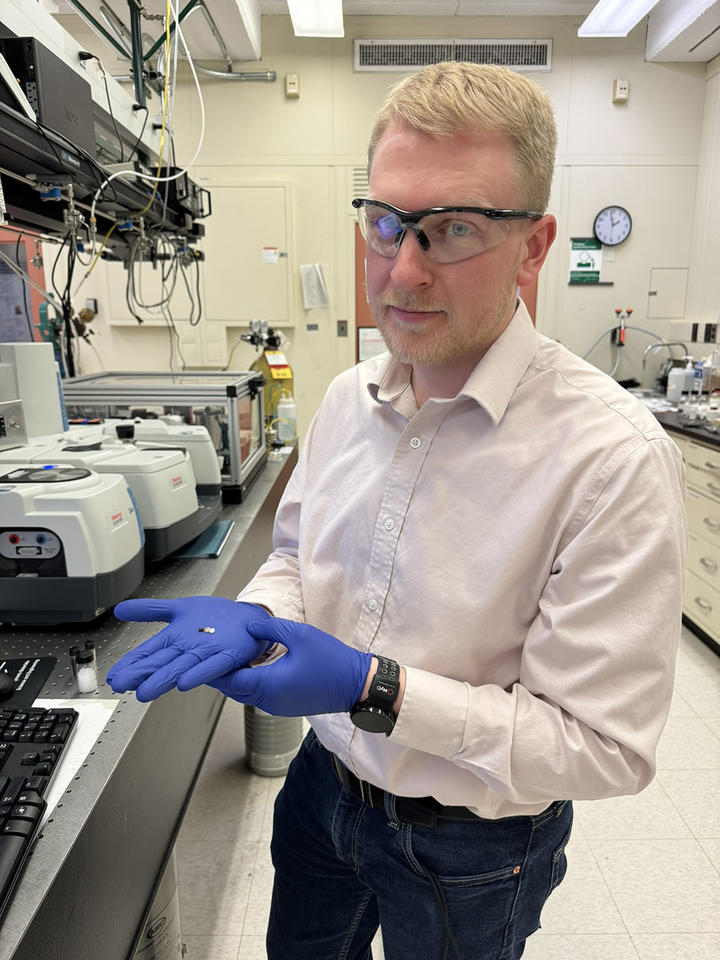 Brad Sutliff displays small circles of plastic on his gloved palm as he stands in the lab wearing safety glasses. 