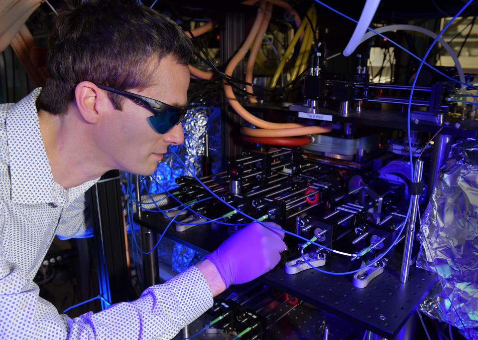 Stephen Eckel wears dark safety glasses as he peers into the complex CAVS apparatus in a darkened lab. 