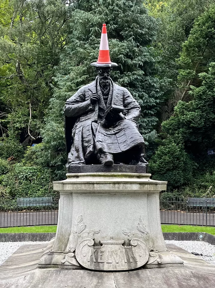 A statue of Lord Kelvin, seated with a notebook, stands in a leafy park. An orange traffic cone is placed on his head like a hat.  